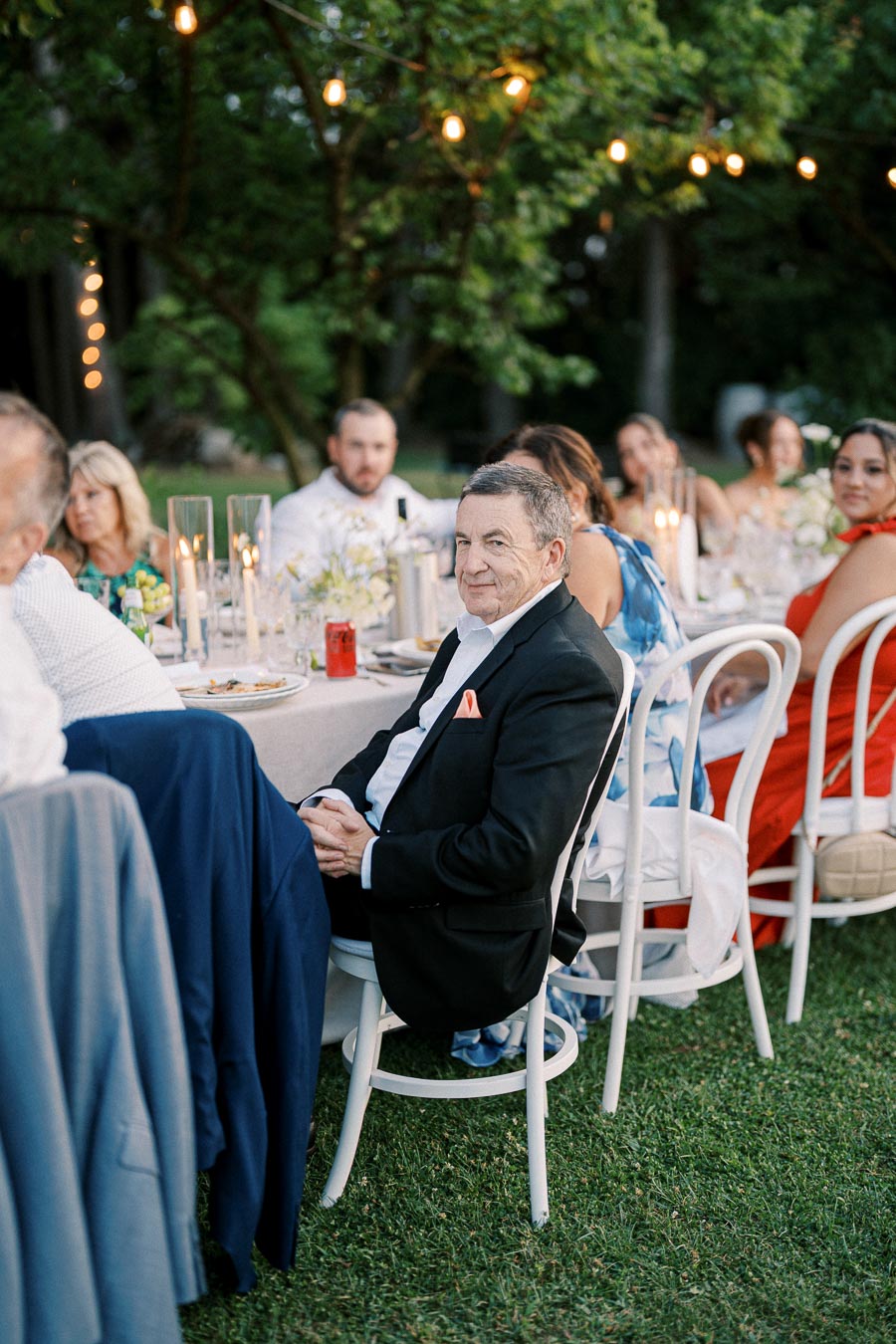 Outdoor garden party with elegantly dressed guests seated at a table adorned with candles and flowers, creating a warm, inviting atmosphere.