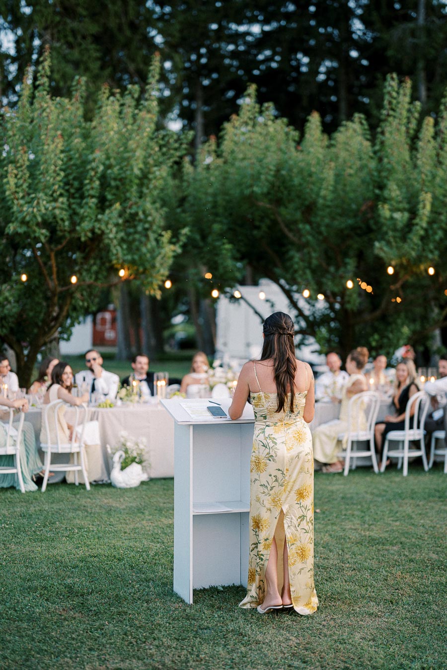 A woman in a floral yellow dress gives a speech at an outdoor wedding reception with guests seated at decorated tables under string lights, surrounded by lush greenery.