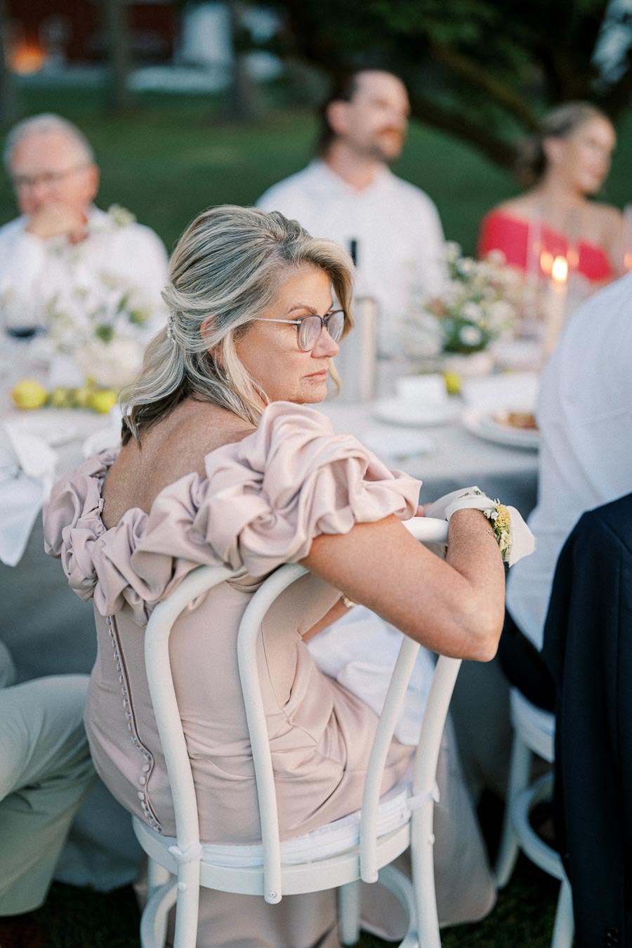 Outdoor evening event with elegantly dressed woman in a light pink ruffled dress sitting on a white chair, surrounded by a beautifully set dinner table with floral decorations and guests.