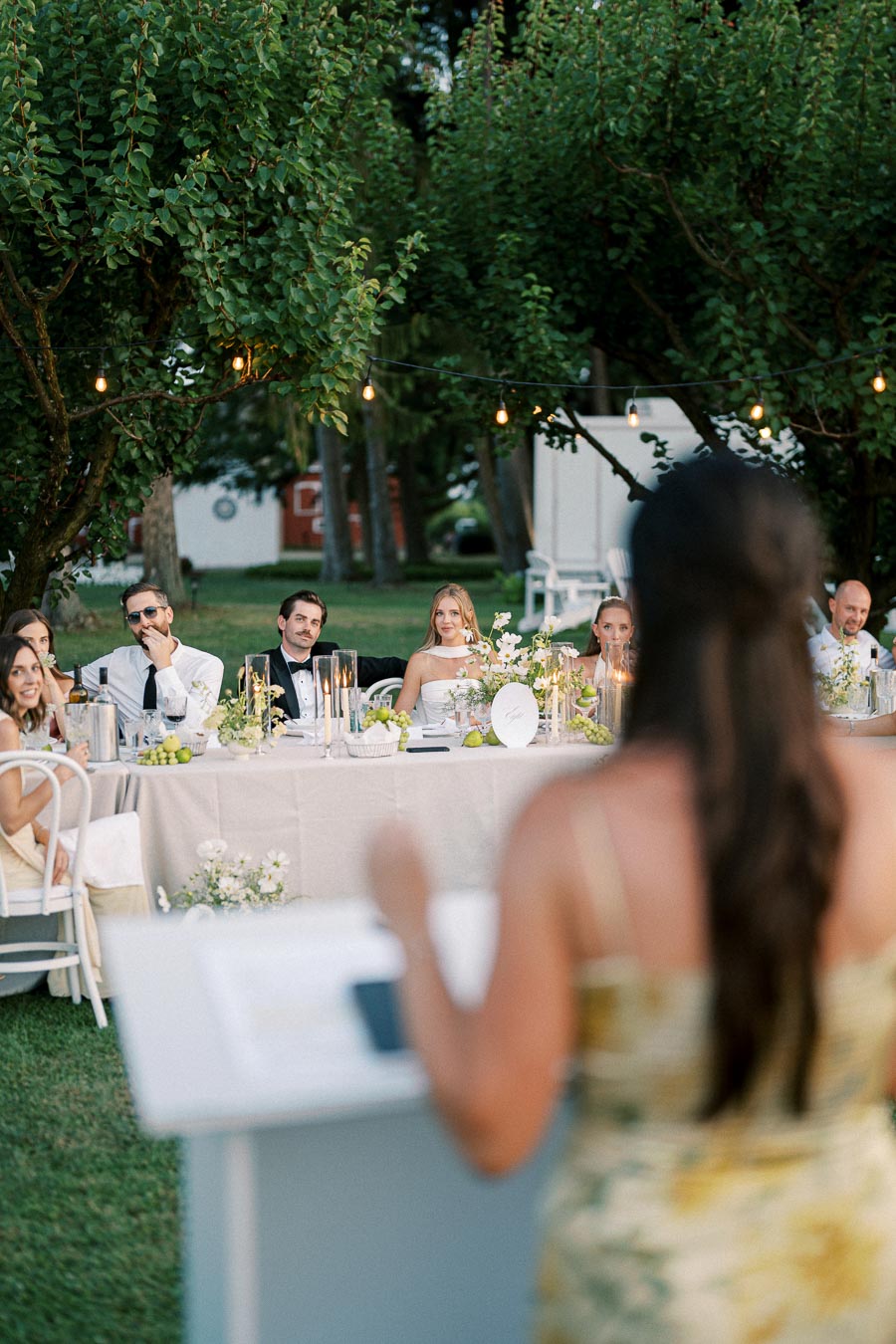 Outdoor wedding reception with guests seated at an elegantly decorated table, listening to a speech. Fairy lights are strung overhead, creating a warm and festive ambiance amidst lush greenery.