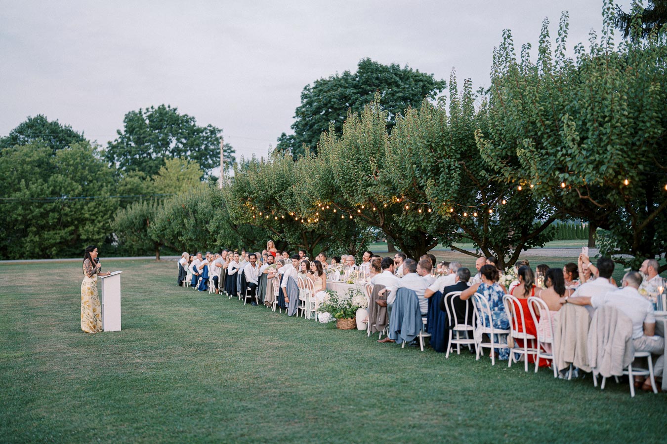 Outdoor wedding reception with guests seated at a long table beneath trees adorned with string lights, focused on a woman giving a speech at a podium.