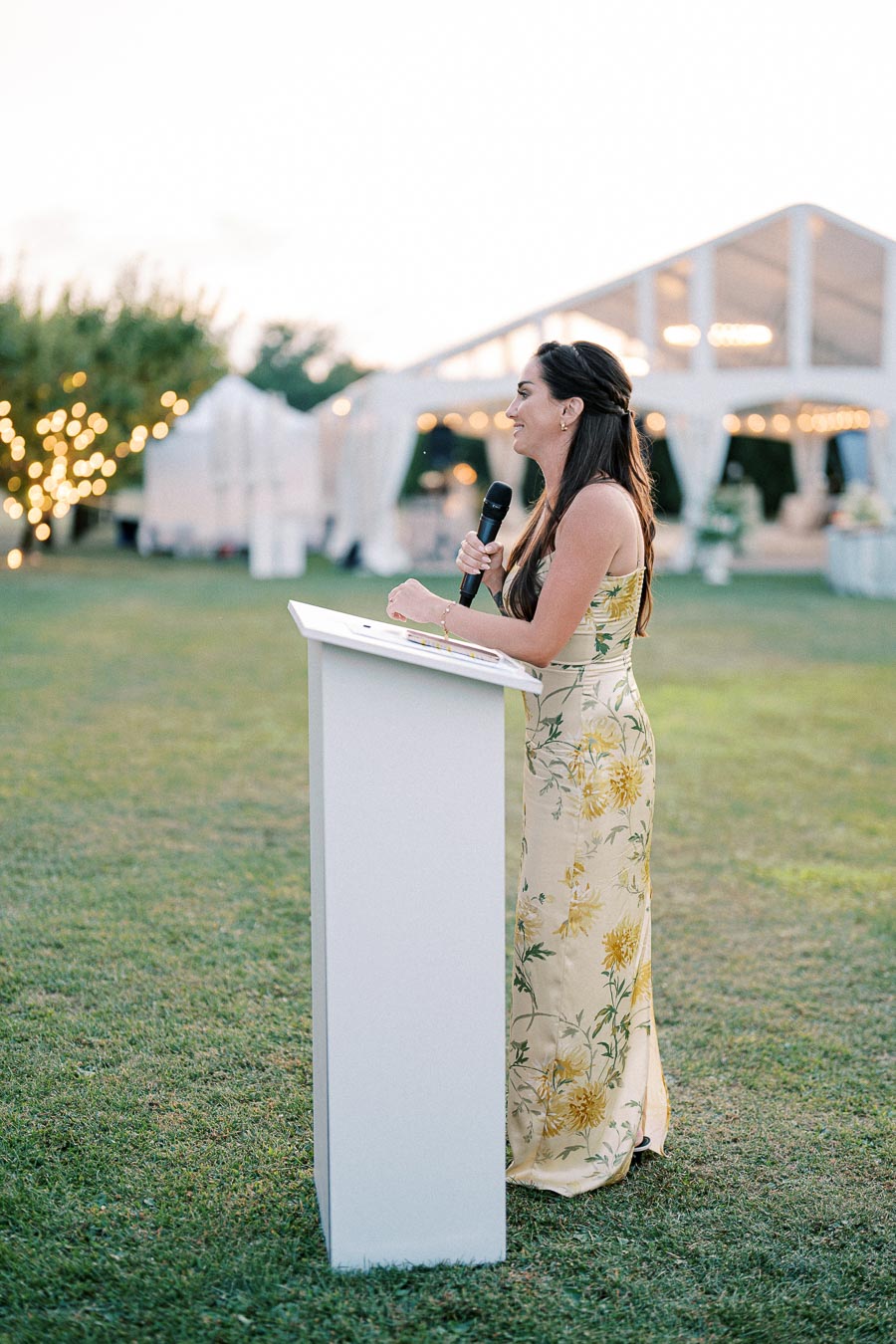 A woman in a floral dress giving a speech at an outdoor wedding reception, standing at a white podium with a microphone, festive lights and a white tent in the background.