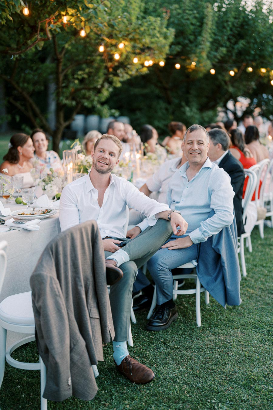 Outdoor evening event with guests seated at a long table adorned with floral arrangements and string lights. Two men in formal attire are smiling at the camera, set in a garden ambiance.