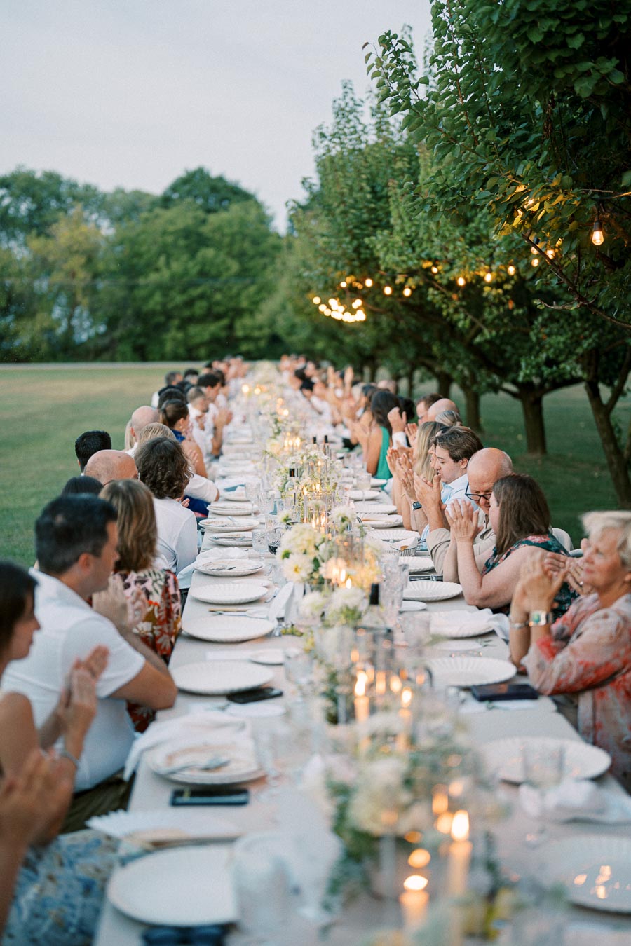 Outdoor dinner party with guests seated at a long, elegantly set table adorned with white flowers and candles, surrounded by lush greenery and twinkling string lights.