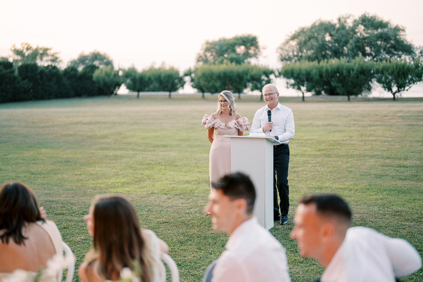 Couple giving a speech outdoors at a formal event, with green lawn and trees in the background, and seated guests listening attentively, captured during a sunny day.