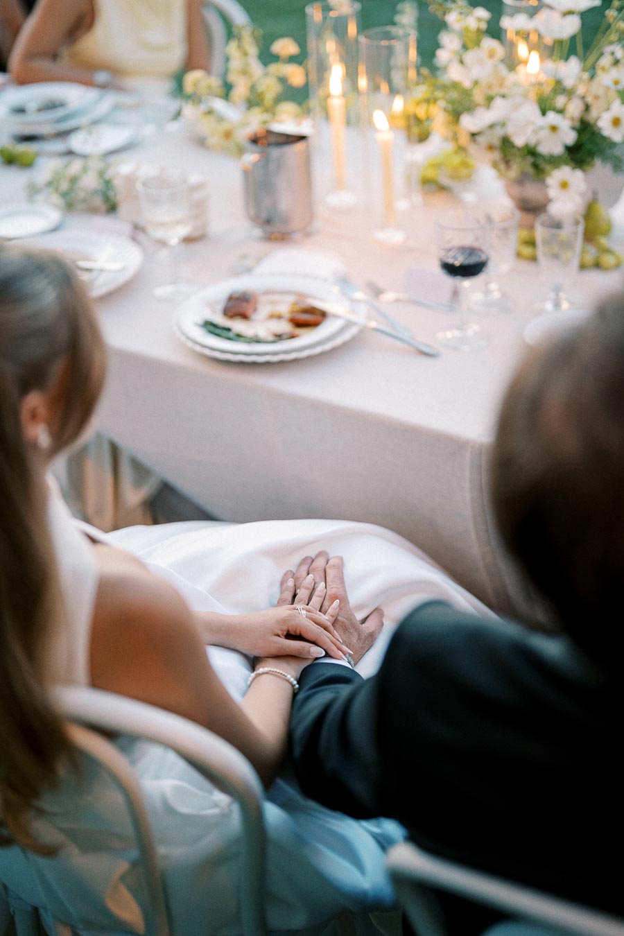Romantic wedding dinner with couple holding hands, seated at elegantly decorated table with candles and flowers, creating an intimate ambiance.
