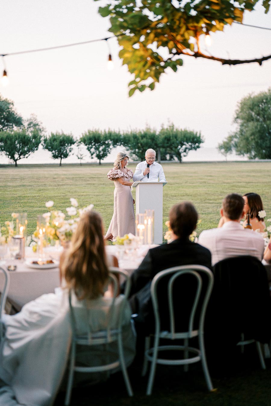 A couple giving a speech at an outdoor wedding reception, surrounded by trees and guests seated at a table with elegant decor and candlelight.