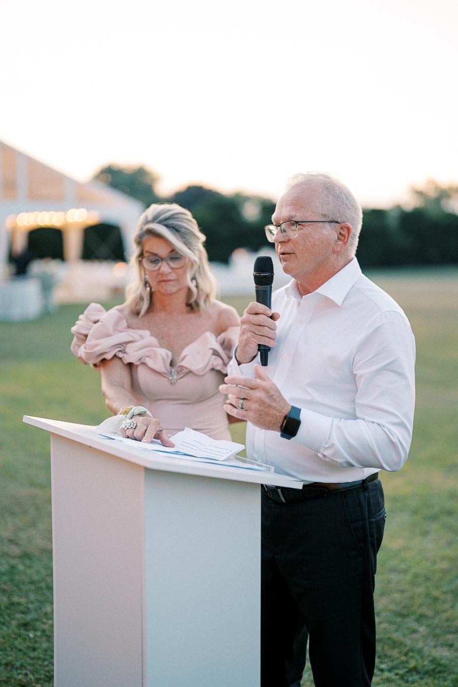 Man speaking into a microphone at an outdoor event, standing at a podium beside a woman in a formal dress, with a soft-focus background.