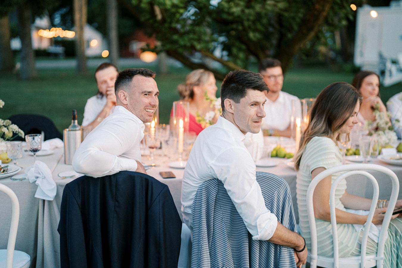 Outdoor dinner party with guests laughing and enjoying a candlelit table setting, featuring elegant white chairs and a green, tree-filled background.