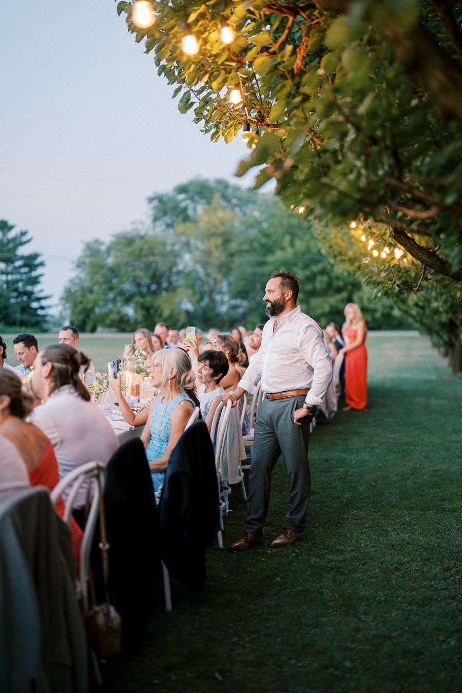 Outdoor evening wedding reception with guests seated at a long table under string lights, surrounded by greenery, and a standing man in a white shirt enjoying the event.