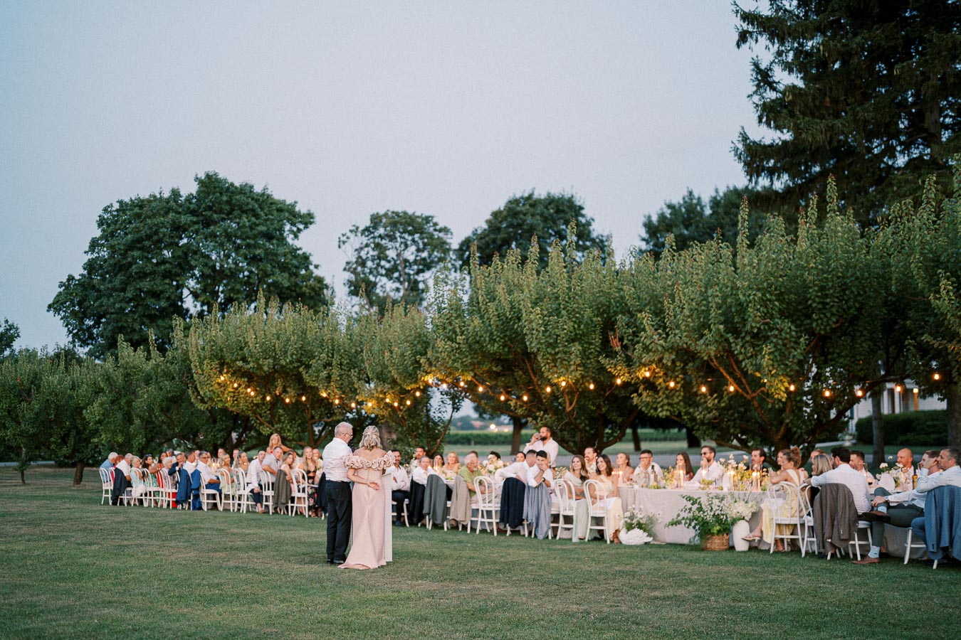 Outdoor evening wedding reception with a crowd of guests seated at elegantly decorated tables under string lights, surrounded by lush greenery, as a couple dances on the lawn.