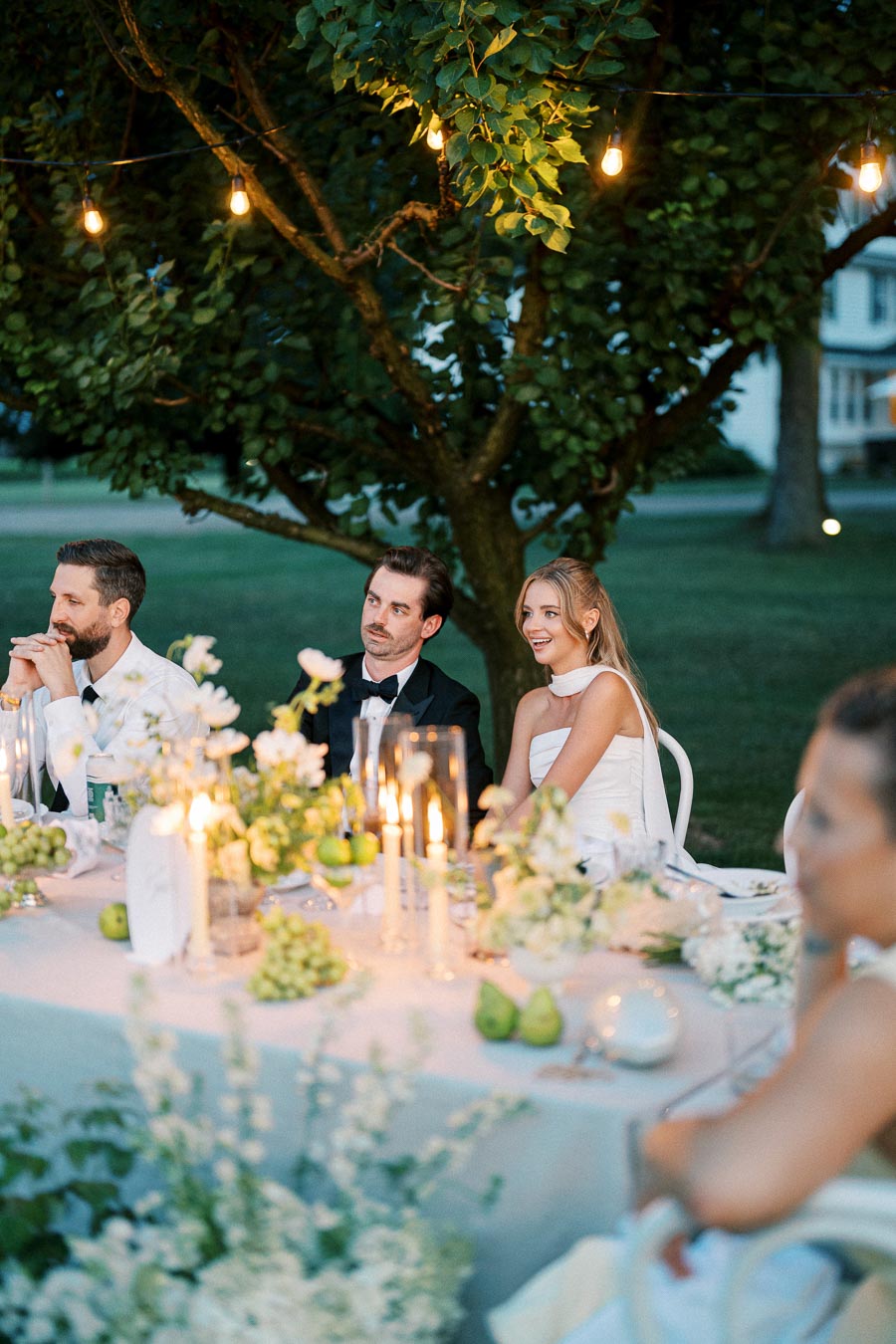 Elegant outdoor evening wedding reception with guests sitting at a beautifully decorated table adorned with white flowers, candles, and string lights under a lush tree.