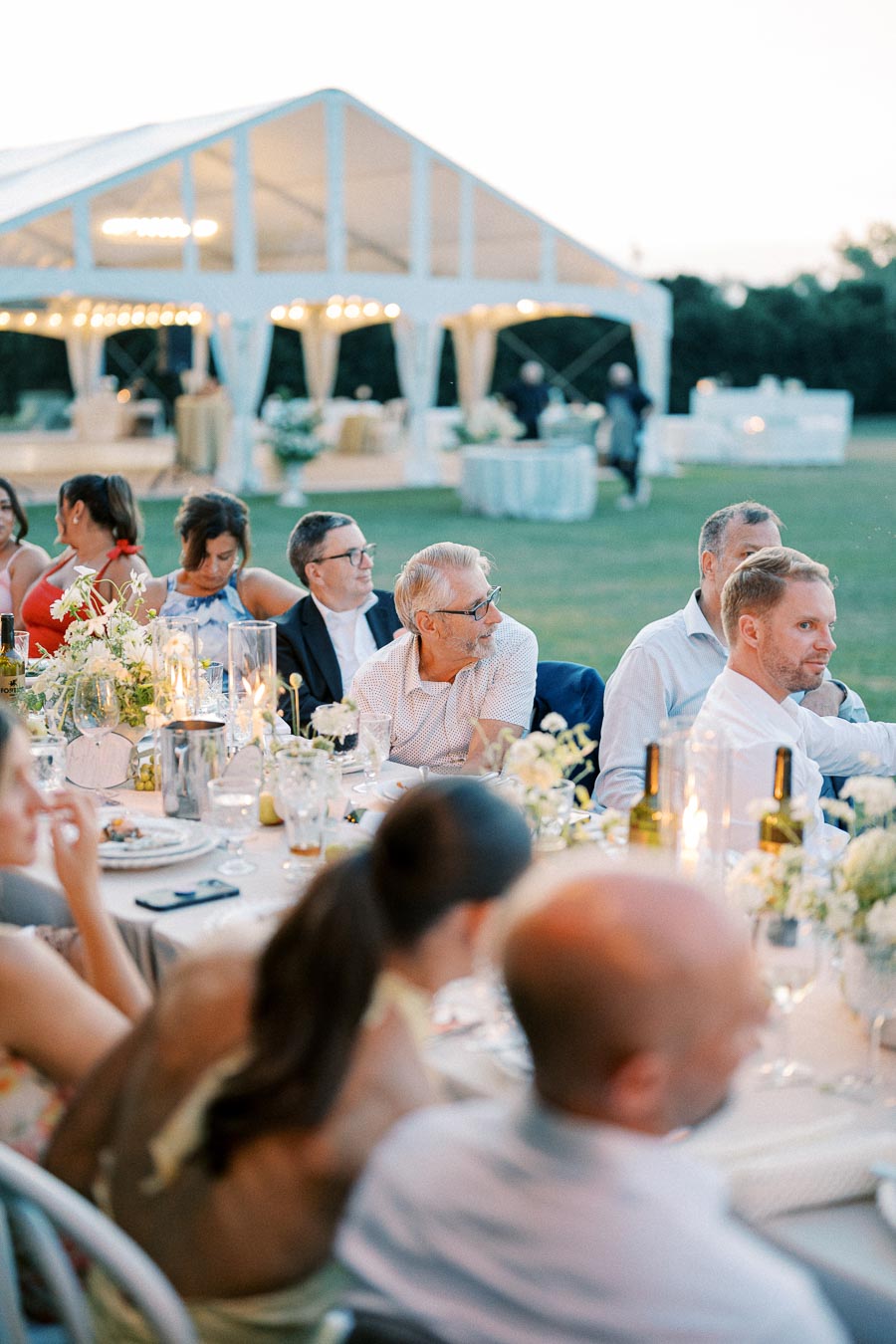 Guests enjoying an outdoor wedding reception under a white tent, with elegantly set tables featuring floral centerpieces and candlelight.