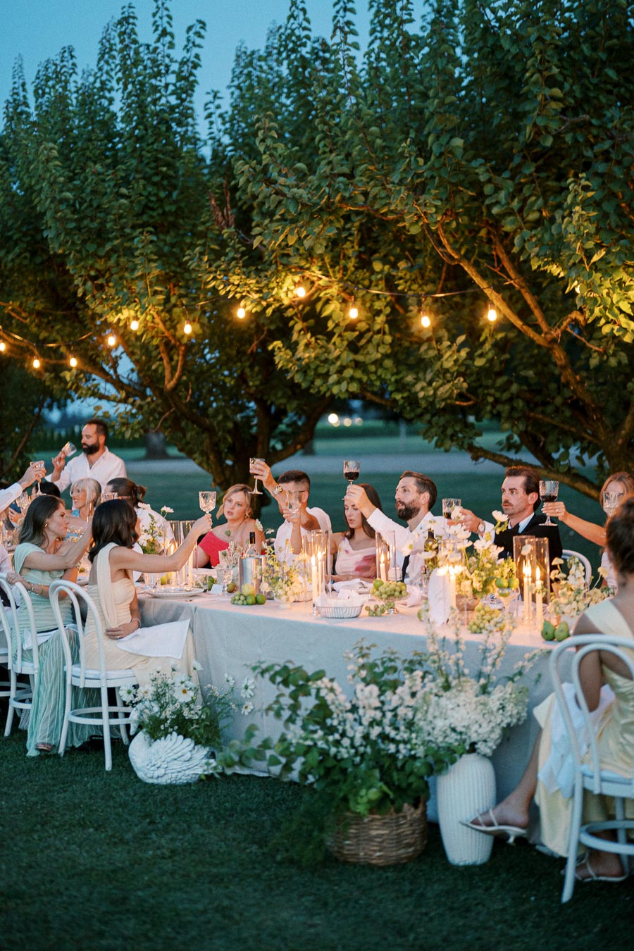Outdoor garden party dinner scene with guests raising glasses for a toast at a beautifully decorated table under string lights in the evening.