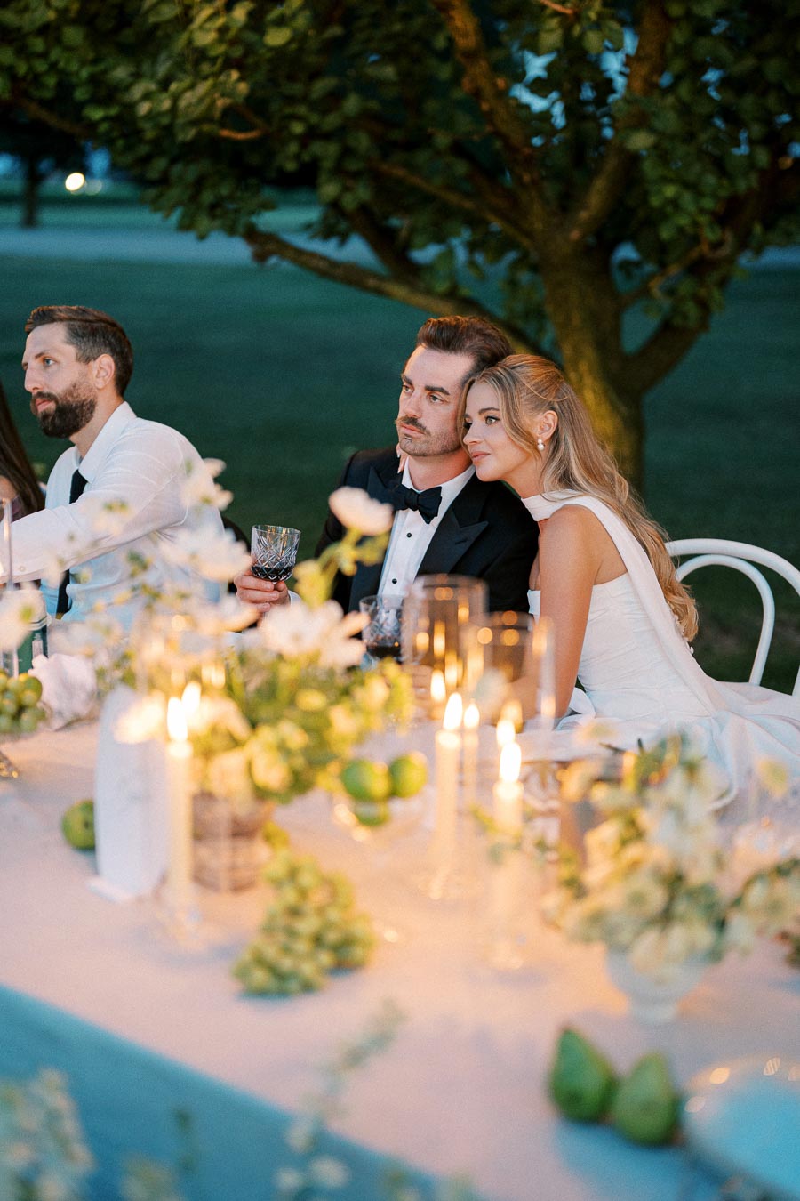 Elegant outdoor wedding dinner setting with a couple in formal attire seated at a candlelit table, adorned with white flowers and green apples, under a tree in a serene evening ambiance.