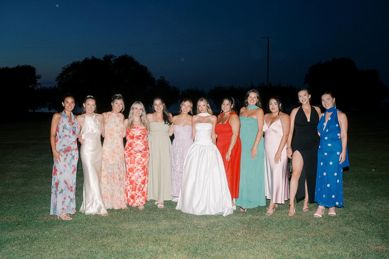 A group of women in elegant dresses standing together on a lawn in the evening, featuring a bride in a white gown surrounded by bridesmaids in colorful attire.