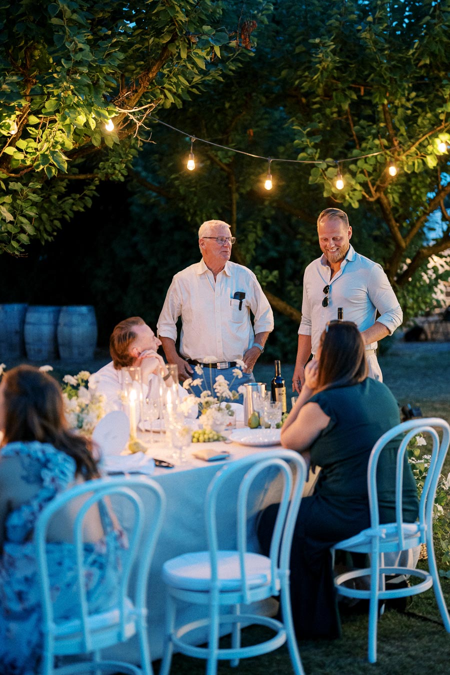 Outdoor evening dinner party with people gathered around a table under string lights, surrounded by greenery.