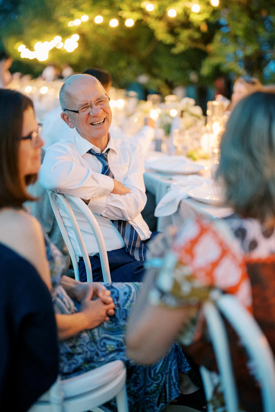 A joyful elderly man laughing and enjoying an outdoor evening event, surrounded by warmly lit string lights and seated guests at a festive dinner table.