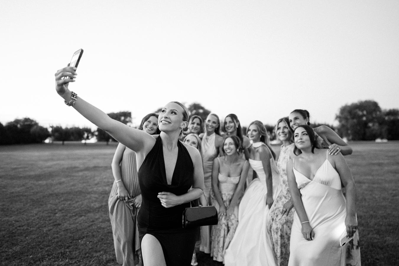Group of women in elegant dresses taking a selfie outdoors in a grassy field during sunset, celebrating an event together.