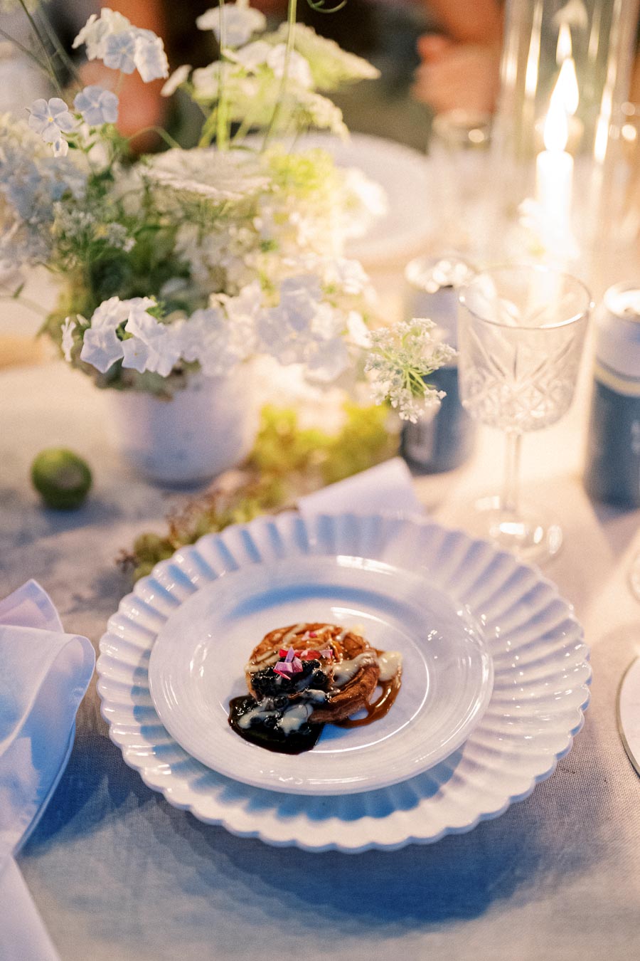 Elegant table setting with a gourmet dish on a decorative white plate, surrounded by white floral arrangements and soft candlelight.