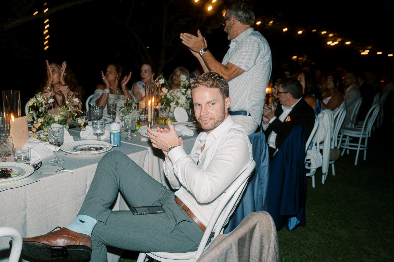 A man in a white shirt sits at an elegantly set dinner table with other guests, clapping during a nighttime outdoor event. The table is adorned with candles and floral arrangements, creating a warm and festive atmosphere.