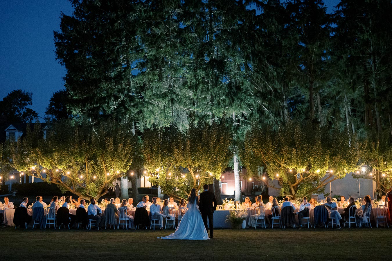 Outdoor evening wedding reception under twinkling string lights, with guests seated at long tables surrounded by lush trees.