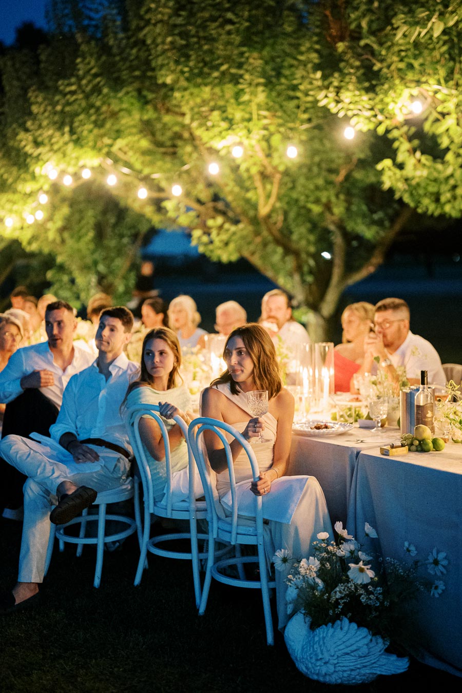 Outdoor dinner party under string lights with guests seated at elegantly decorated tables, enjoying an evening gathering in a lush garden setting.