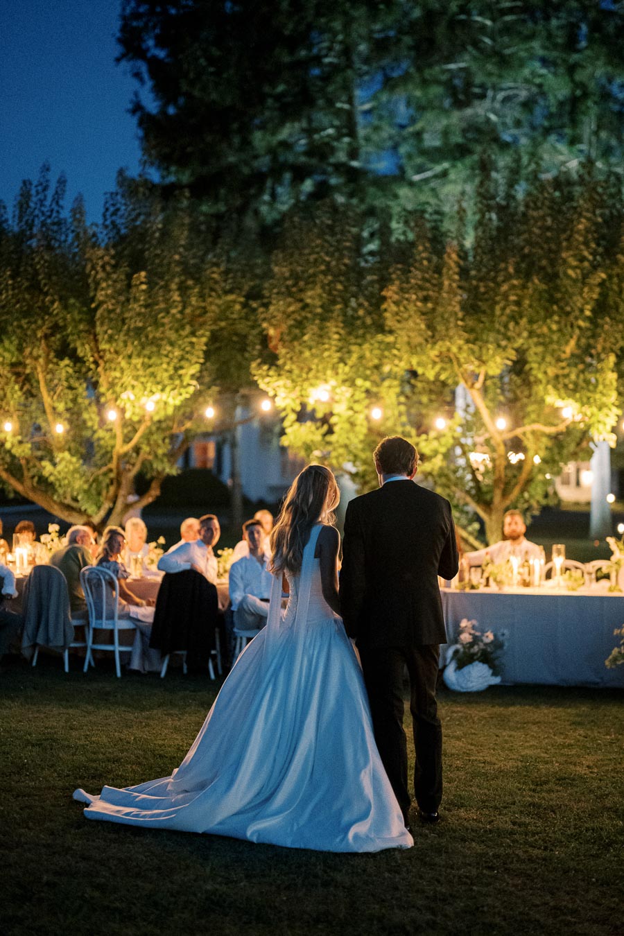 Outdoor evening wedding reception with a bride in a flowing gown and groom standing near a candle-lit table, surrounded by guests under string lights and trees.