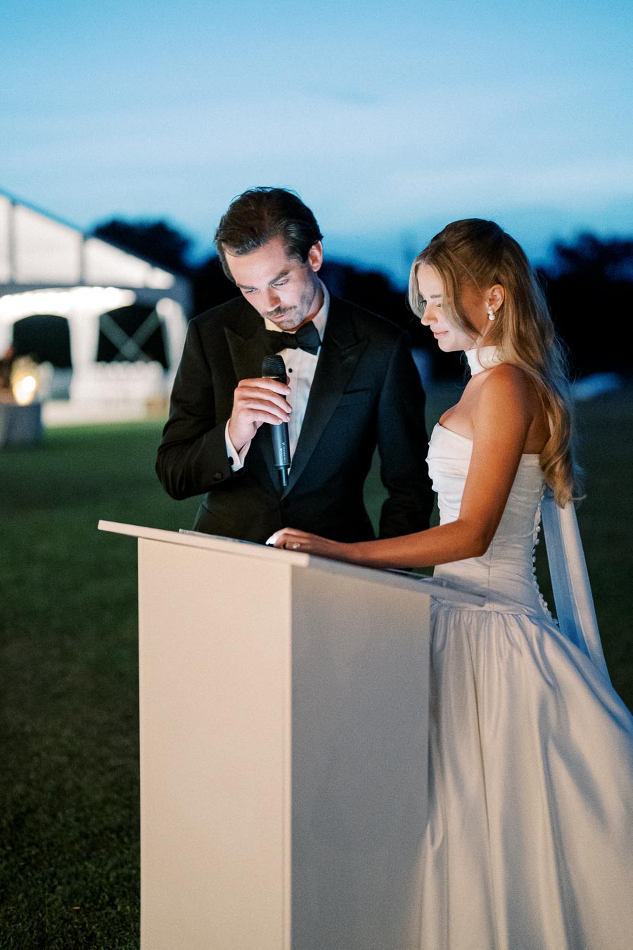 A bride and groom in formal attire giving a speech at their outdoor wedding ceremony during the evening.