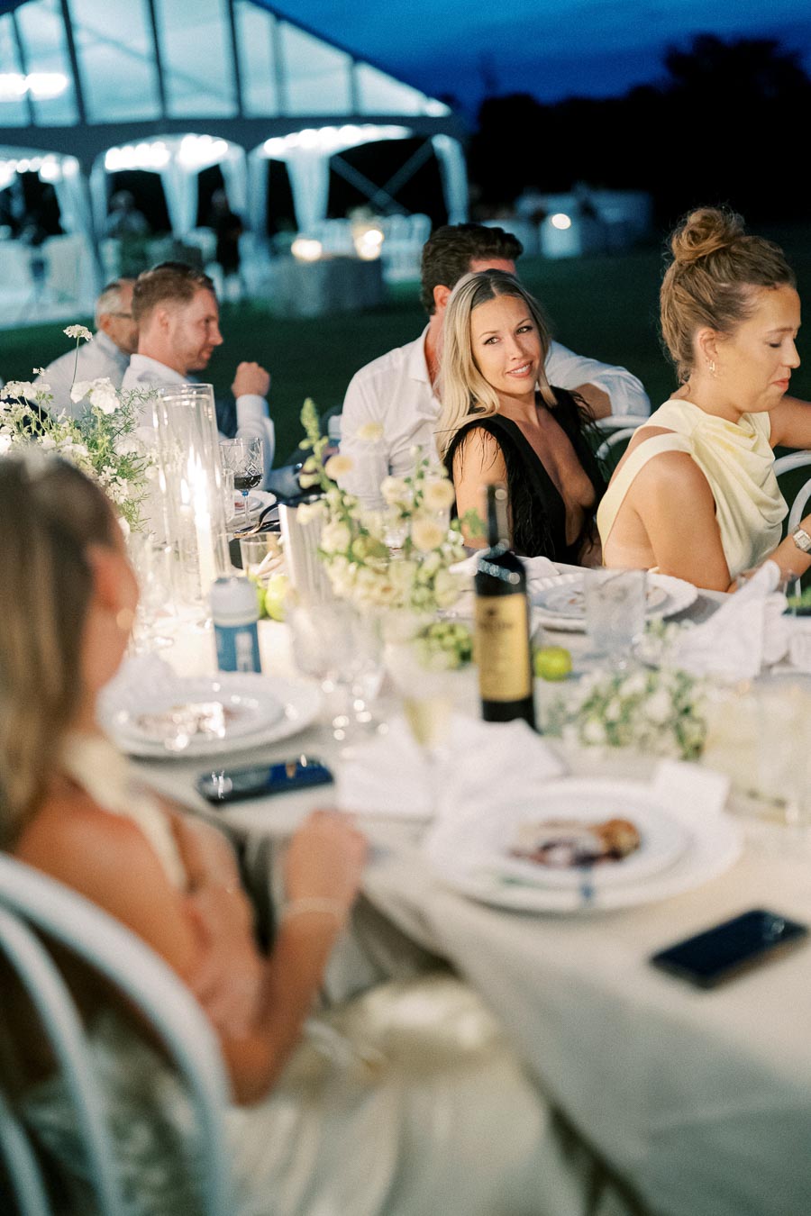 Elegant outdoor evening dinner party under a canopy with guests seated at a beautifully decorated table adorned with flowers and candles.