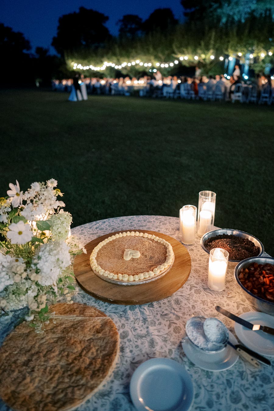 Outdoor evening wedding table setup with rustic pie, candles, and decorative flowers. A reception with string lights and guests is visible in the background, creating a warm, festive atmosphere.