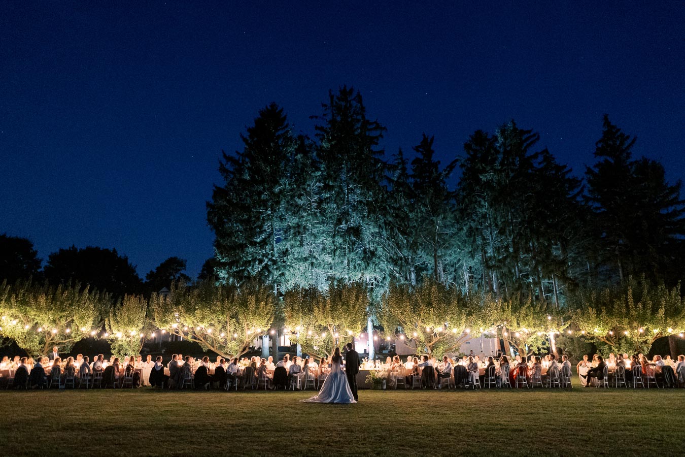 Outdoor evening wedding reception with guests seated at long tables under string lights, with a forest backdrop and a couple standing in the foreground, set against a dark blue sky.