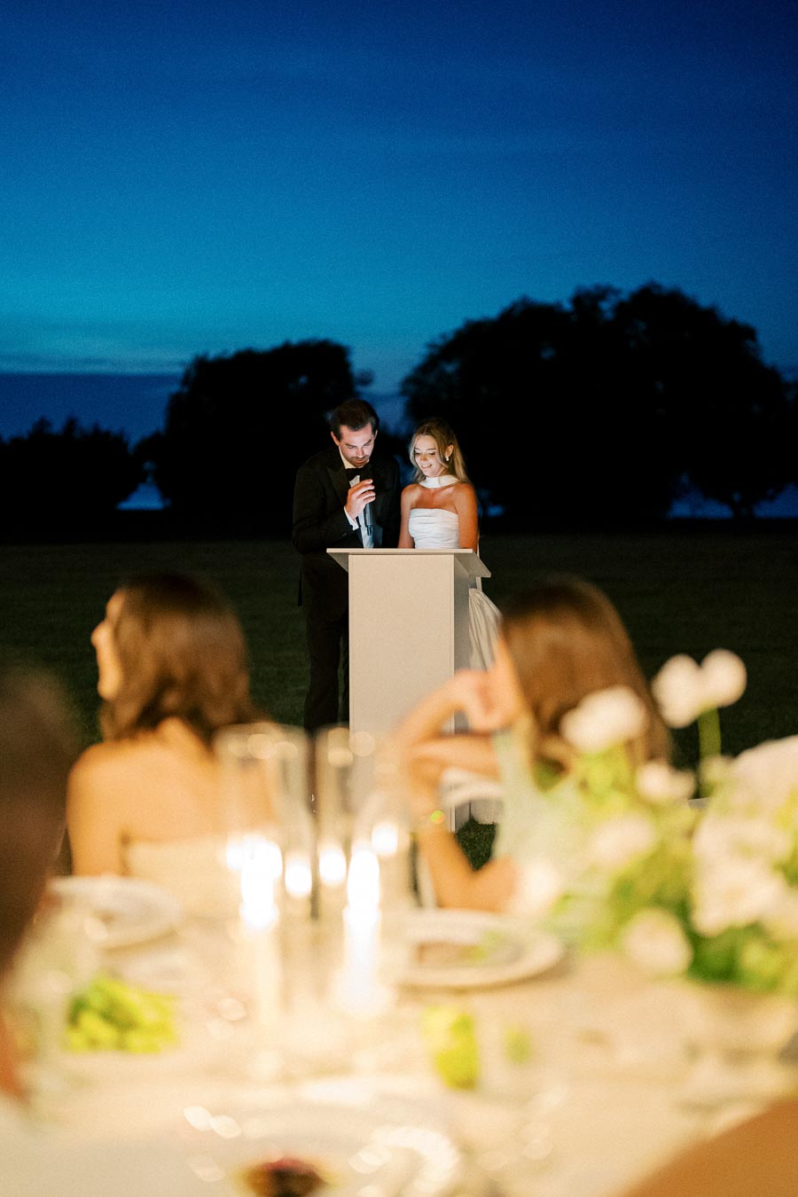 Bride and groom give a heartfelt speech during an outdoor evening wedding reception, with a beautifully decorated table in the foreground and a vibrant blue sky as a backdrop.