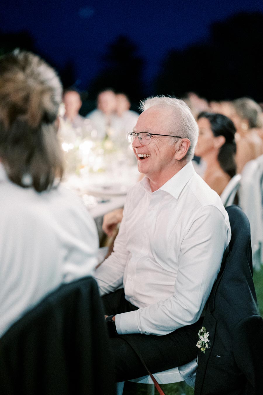 A joyful man in glasses and a white shirt laughing at an outdoor evening event, surrounded by other people at a beautifully lit table.