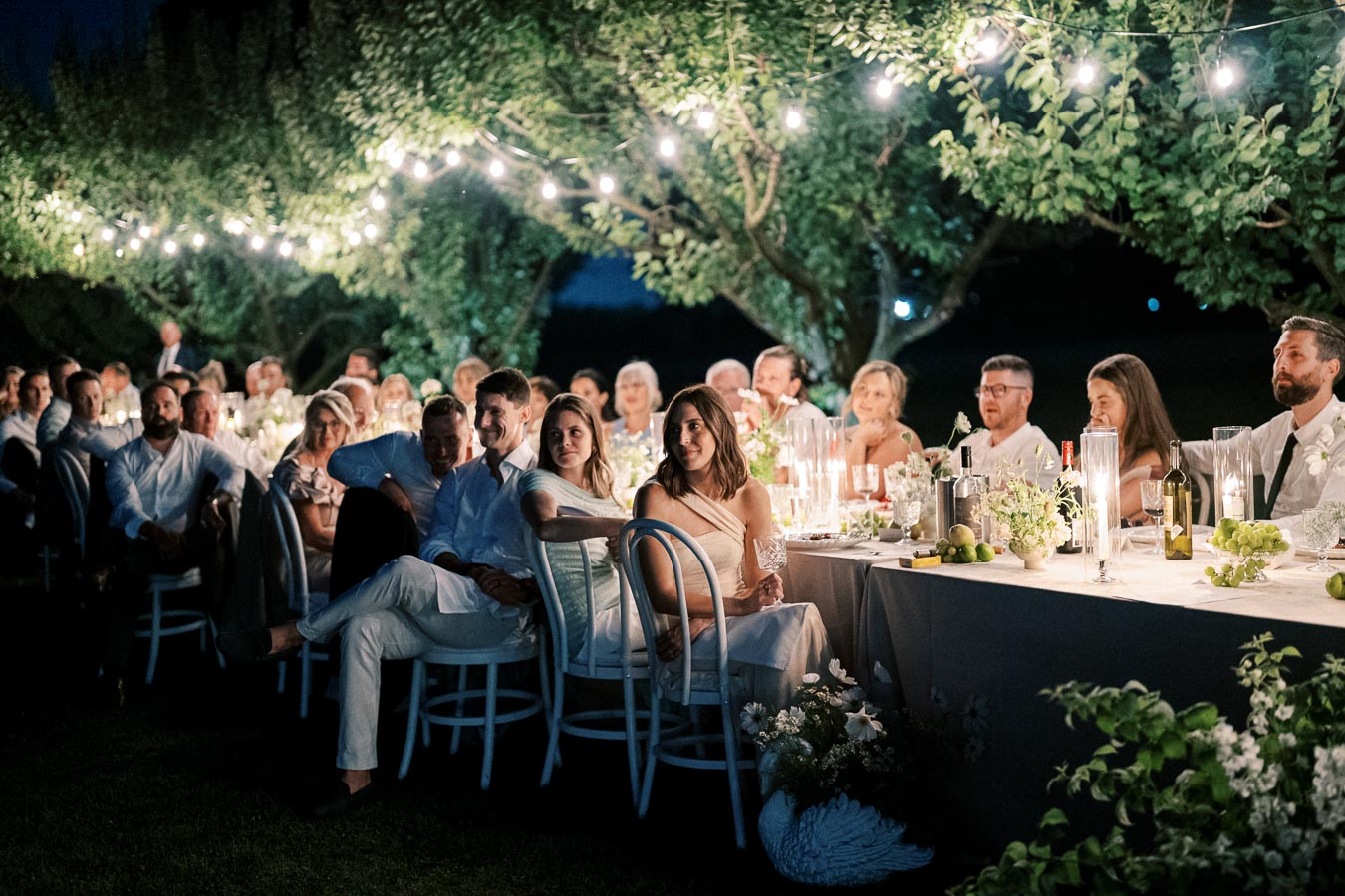 Outdoor evening wedding reception with guests seated at a long table under string lights, surrounded by lush greenery and elegantly set with candles and flowers.