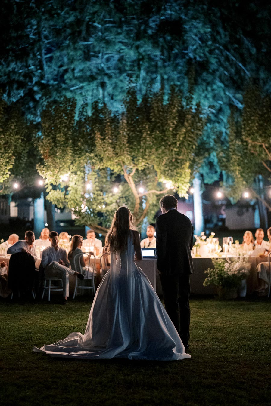 Outdoor evening wedding reception with a bride in a white gown and a groom in a suit standing together, addressing seated guests under string lights and surrounded by trees.