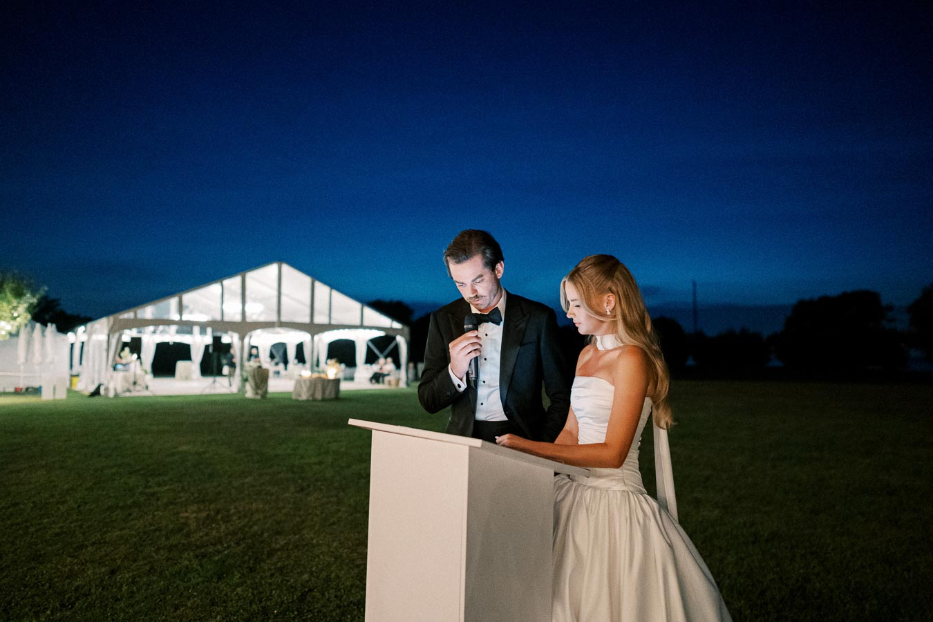 Outdoor wedding reception with a bride in a white gown and groom in a tuxedo delivering a speech under a starry night sky, with a white tent in the background.