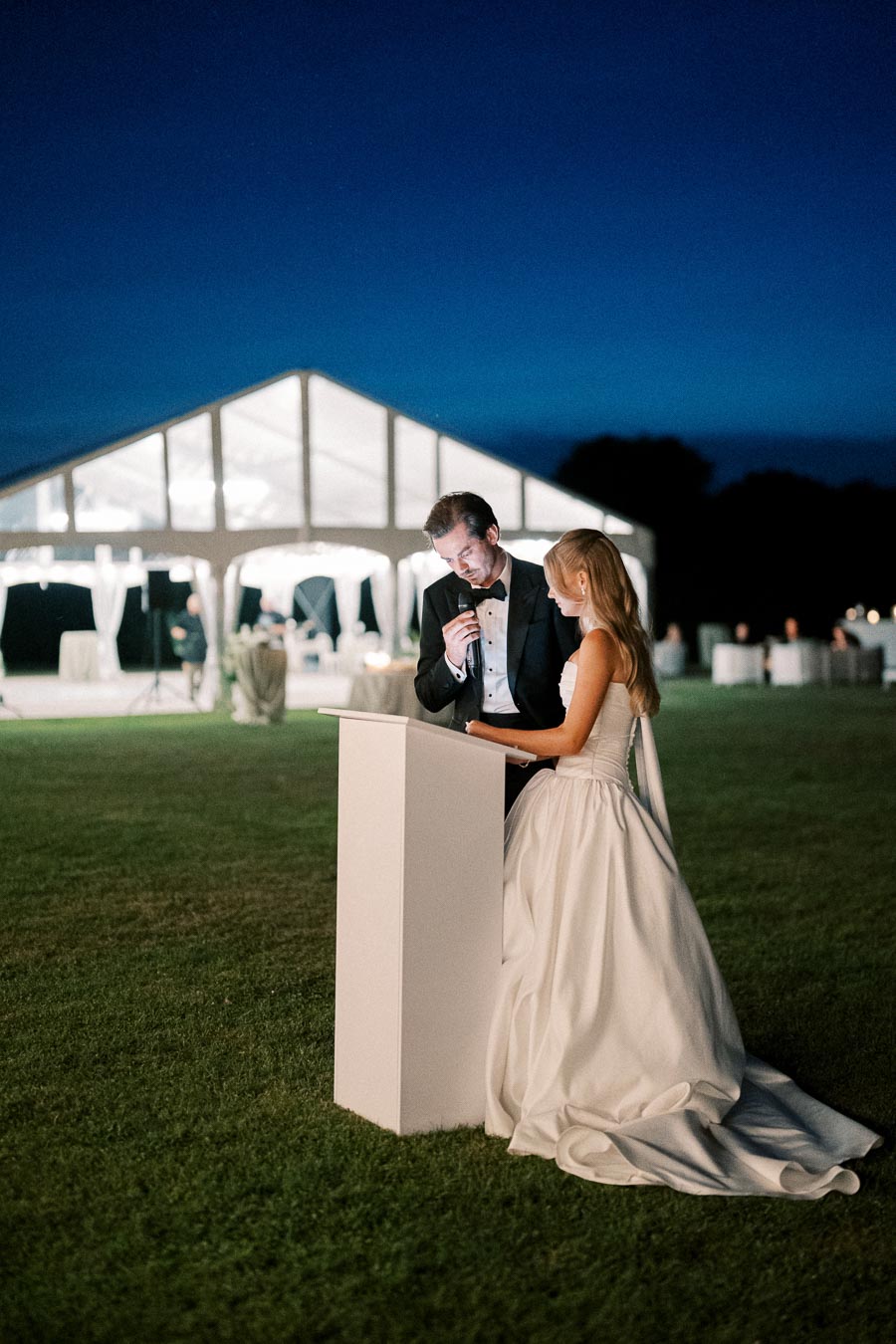 A bride and groom deliver a speech during their outdoor wedding reception at night, standing at a podium in front of a beautifully lit tent.