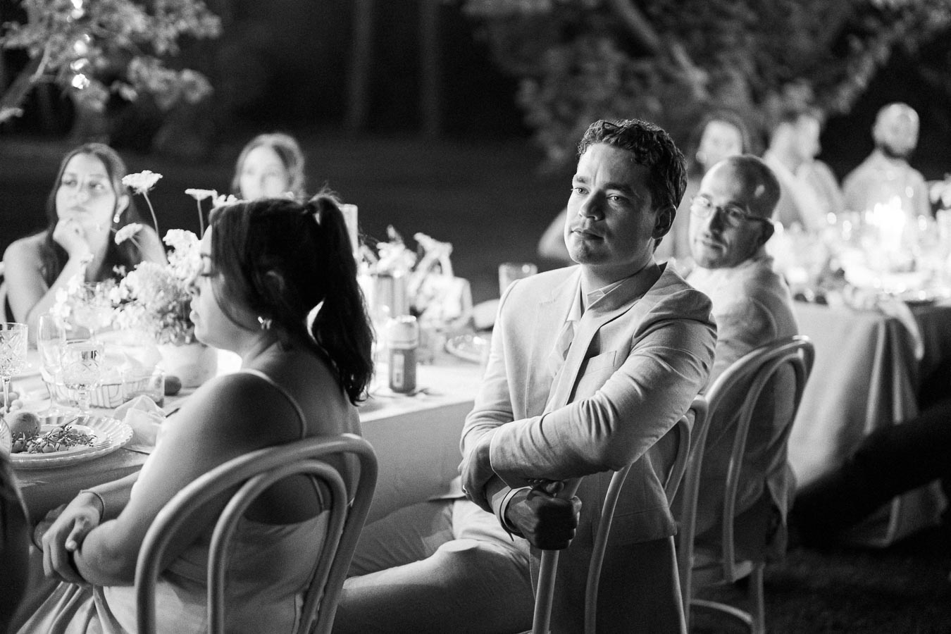 Black and white photo of a group of people seated at a table during an outdoor event, with a man in a light suit prominently in focus.