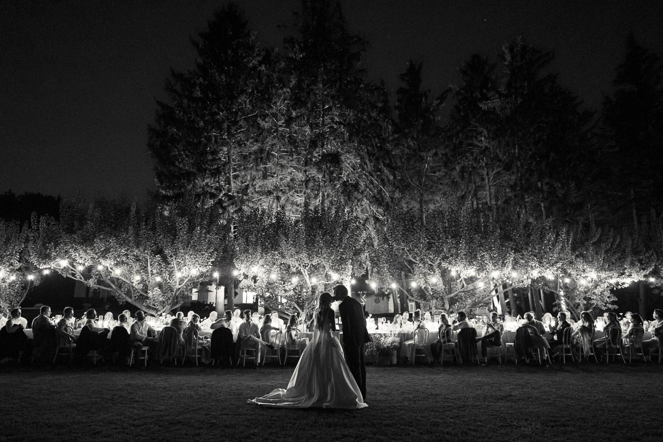 A black and white image of a romantic outdoor wedding reception at night, featuring a bride and groom sharing a moment in the foreground with guests seated at a long table under string lights and trees.