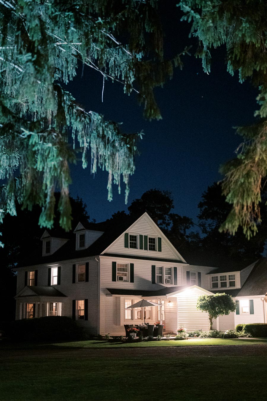 Beautiful night view of a charming white colonial-style house with green shutters, illuminated by warm outdoor lighting, surrounded by leafy trees.