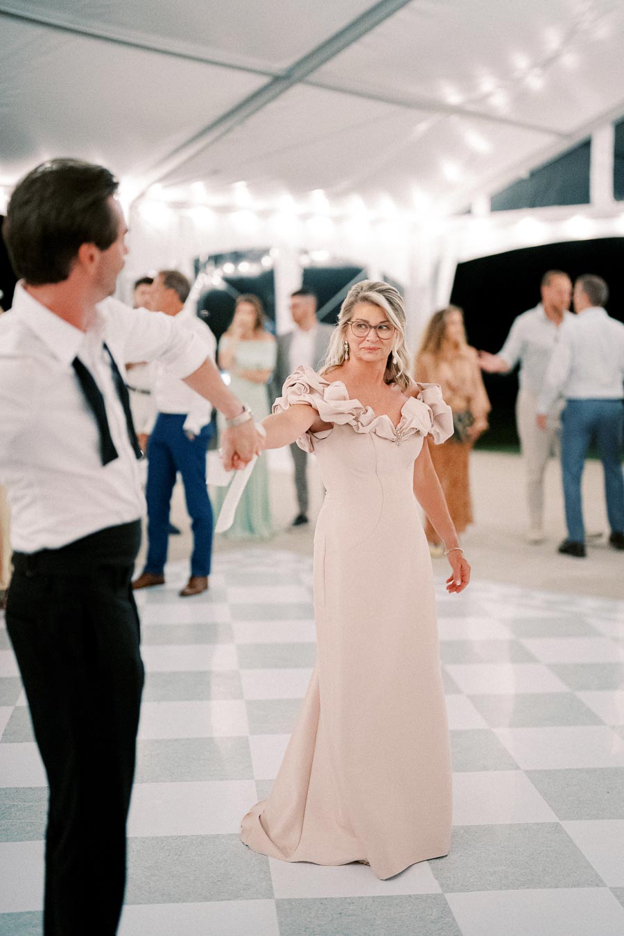 A woman in a flowing pastel pink gown dances with a man in a white shirt and black tie on a checkered dance floor under a white tent adorned with string lights, surrounded by elegantly dressed guests.