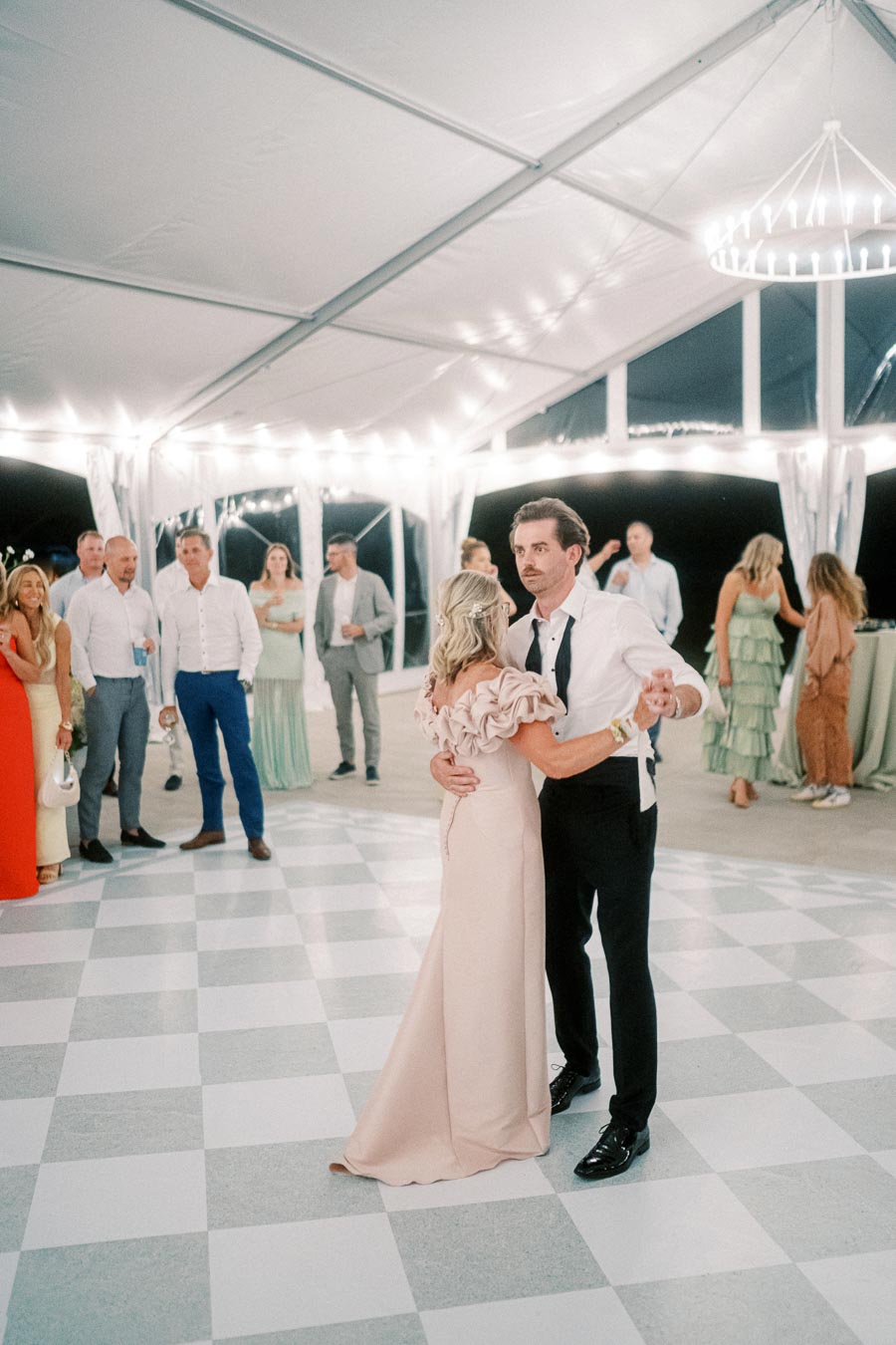 A couple elegantly dances on a checkered floor under a beautifully lit tent, surrounded by well-dressed guests at a formal event.