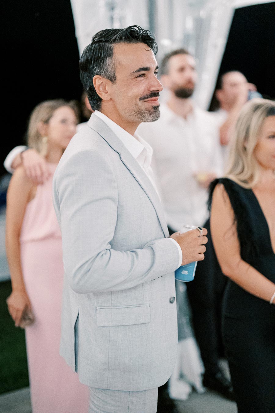 Man in light gray suit holding a drink at an elegant event with people in formal attire in the background.