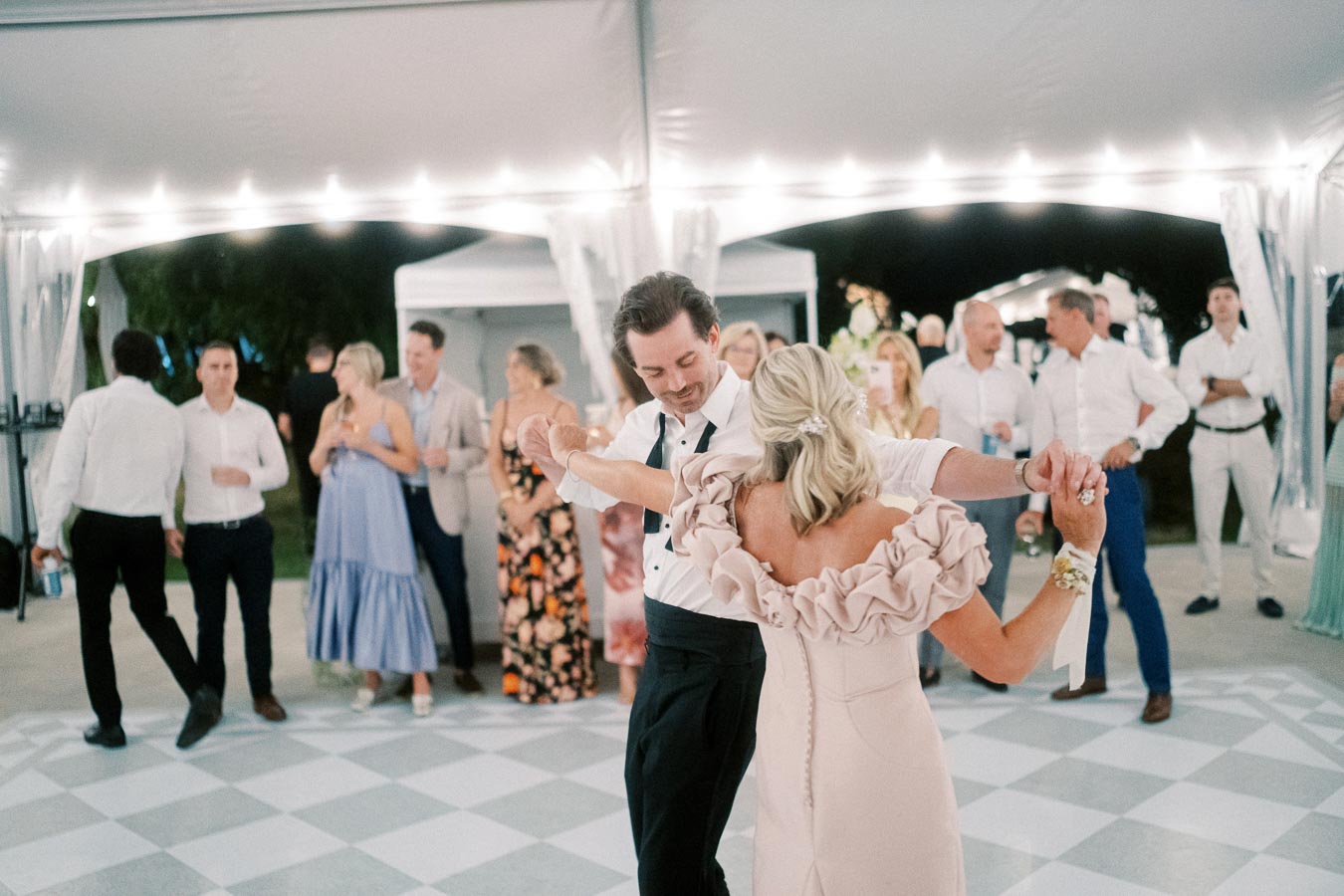 Couple dancing at an outdoor wedding reception, surrounded by elegantly dressed guests under illuminated tent.