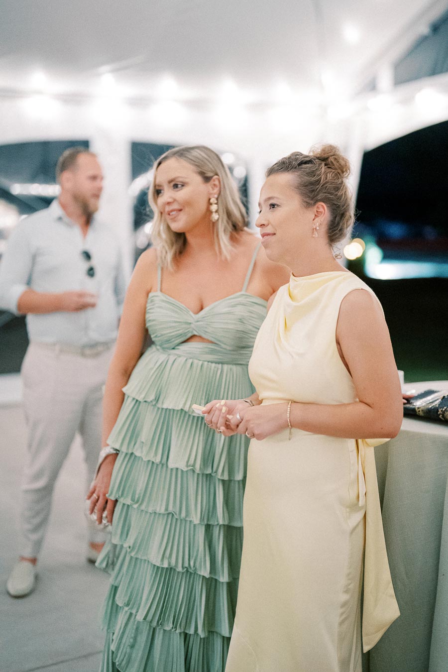 Two women in elegant dresses, one in a layered mint green gown and the other in a draped yellow dress, stand together at a lively event under string lights, with a man slightly blurred in the background.