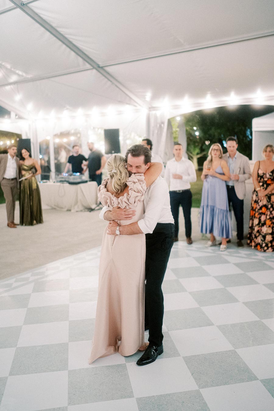 Elegant wedding reception with guests celebrating on the dance floor, featuring a heartfelt embrace under a beautifully lit tent.