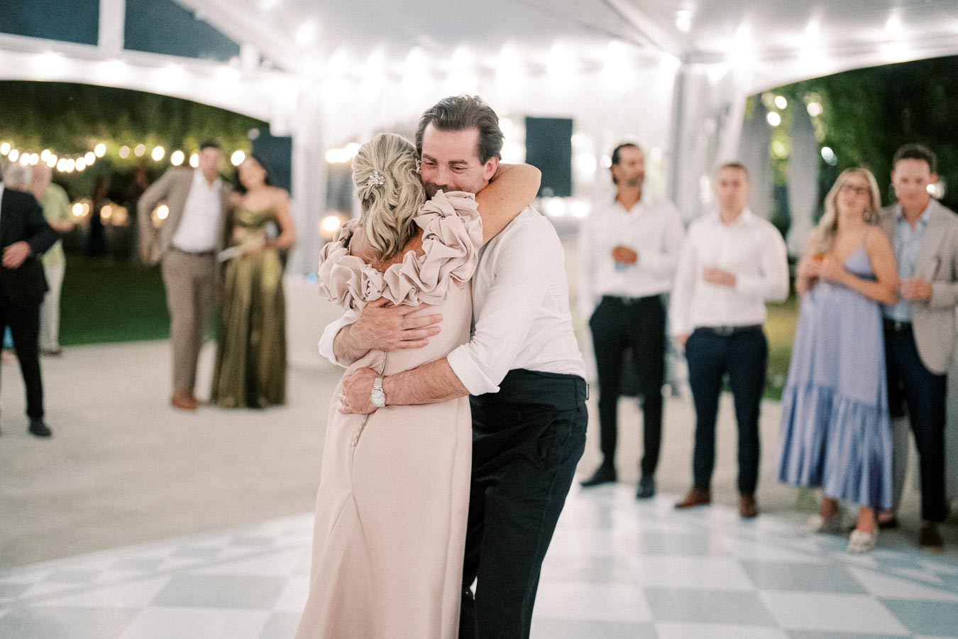 A joyful couple shares a warm hug on the dance floor during an elegantly decorated evening event, surrounded by guests wearing formal attire under soft, ambient lighting.
