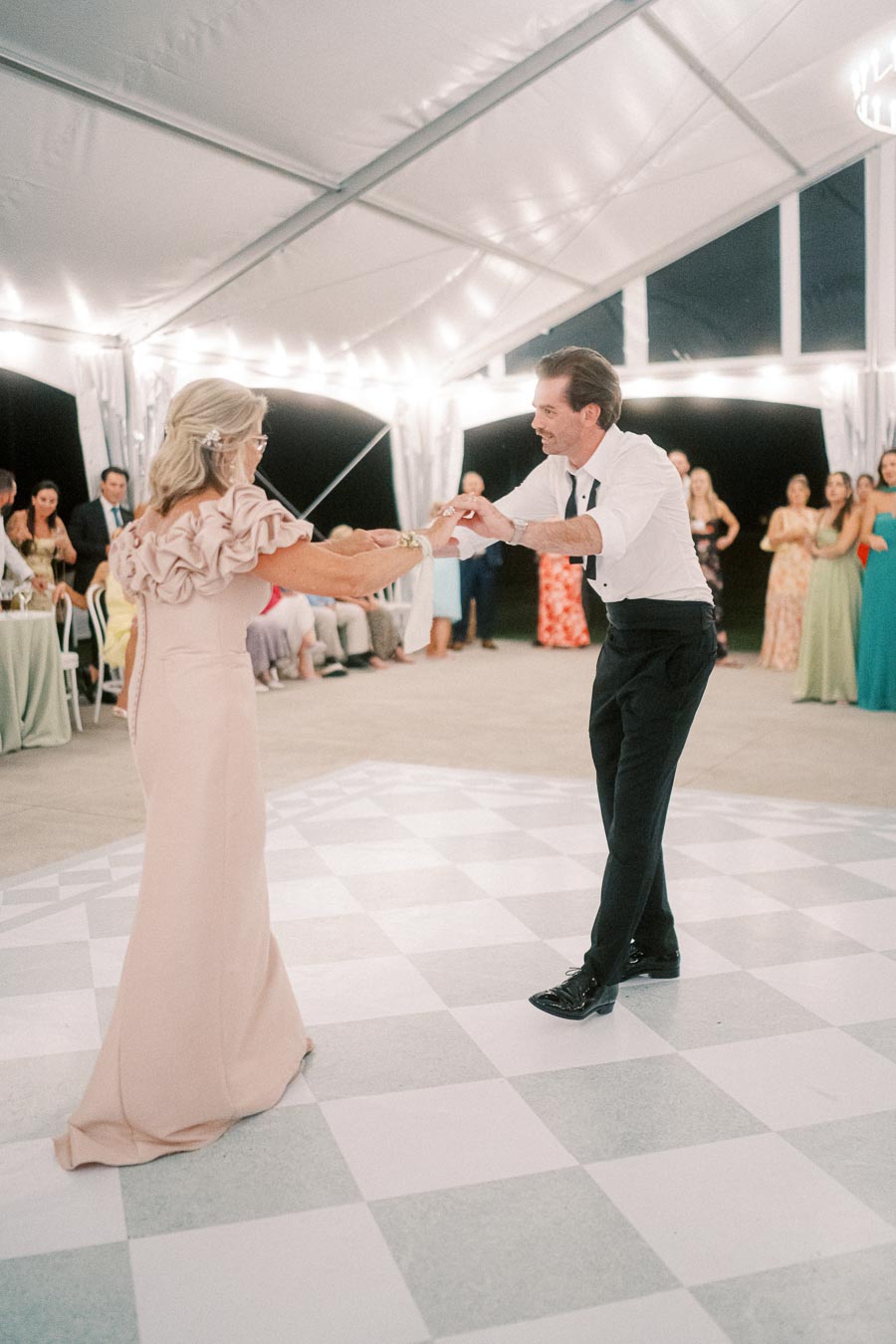 Elegant couple dancing at an outdoor event under a chandelier-lit tent with guests in the background watching.