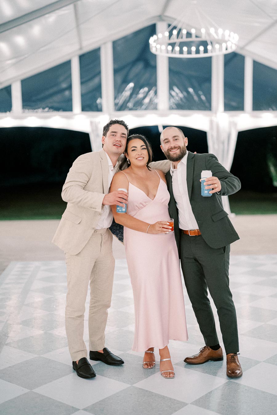 Three people dressed in formal attire, standing closely and smiling at a wedding reception under a white tent with a chandelier.
